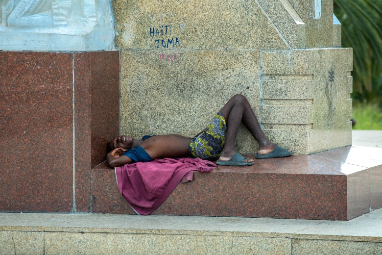 A young man sleeps at the base of the statue of General Petion on November 17, 2022. Right above him, the graffiti writing says Haiti Toma, an expression of fondness for the land. Champ de Mars, in Port-au-Prince, Haiti. Photo by Marvens Compère for The Haitian Times