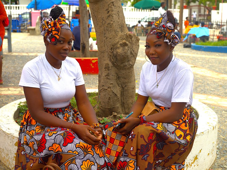 Two women in matching white and brown folkloric attire sit together at Place d’Armes in Cap-Haïtien on May 1, 2025. Photo by Onz Chéry / The Haitian Times.