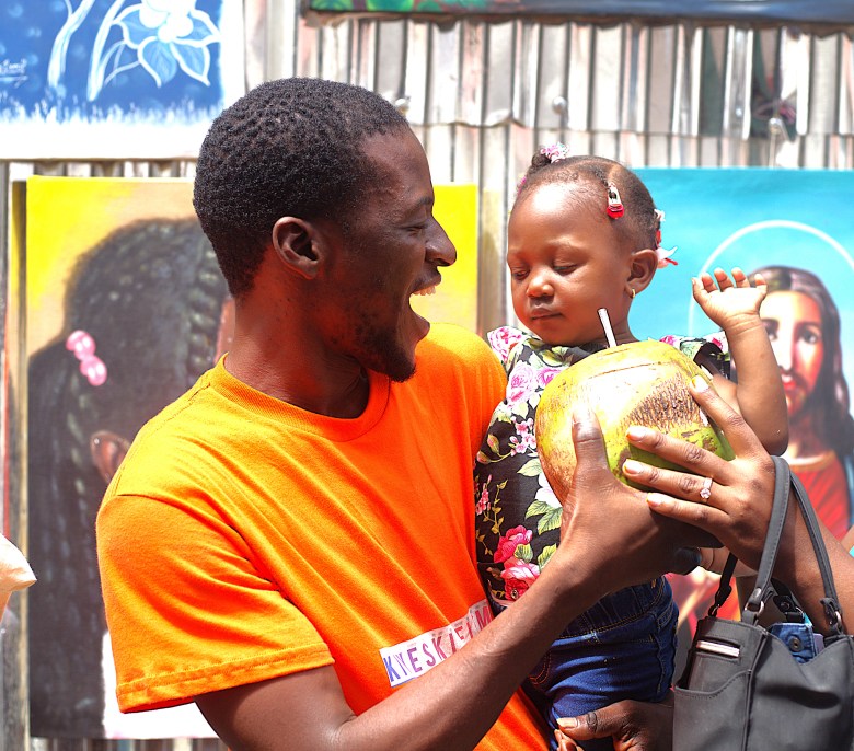 A man helps a baby girl sip from a coconut through a straw at the Institut Sacré-Cœur high school’s food fair on May 1, 2025. Photo by Onz Chéry / The Haitian Times.