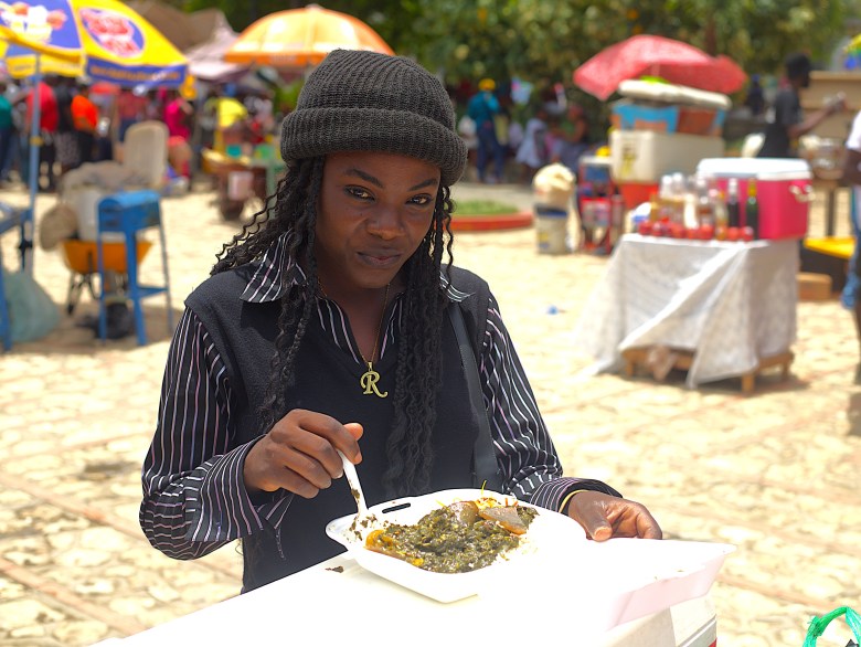 A woman pauses from eating her diri ak lalo to acknowledge the camera at Institut Sacré-Cœur du Cap-Haïtien on May 1, 2025. Photo by Onz Chéry / The Haitian Times.