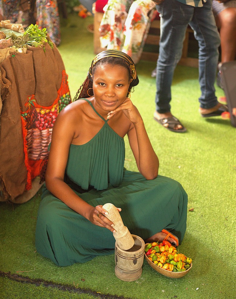 A woman seated on the floor holds a wooden pestle next to peppers during the food fair at Institut Sacré-Cœur du Cap-Haïtien on May 1, 2025. Photo by Onz Chéry / The Haitian Times.