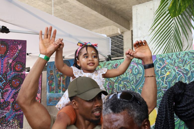 A young girl, accompanied by a relative, enjoys the music of a walking band during Gwoup Eritaj’s Agricultural and Artisanal Resistance Fair in Pétion-Ville, Port-au-Prince, on May 1, 2025. Photo by Dieugo André for The Haitian Times.