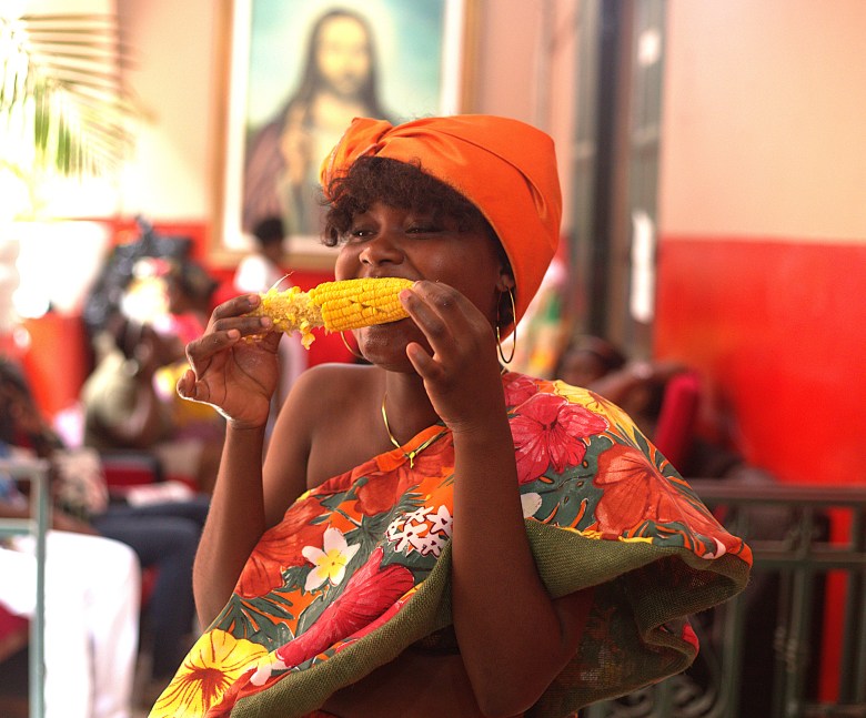 A woman in a red folkloric top eats corn at the food fair at Institut Sacré-Cœur du Cap-Haïtien on May 1, 2025. Photo by Onz Chéry / The Haitian Times.