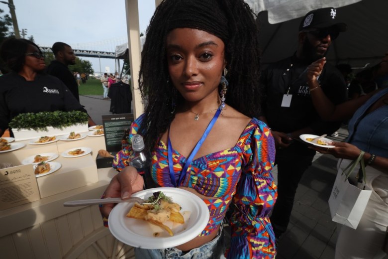 Jessica Merritt from Brooklyn samples lambi pike prepared by Chef Jeffrey Morneau at the Creole Food Festival in Brooklyn on Saturday, Sept. 27, 2025. She described the dish as tangy and fresh. The festival took place at Emily Roebling Plaza under the Brooklyn Bridge. Morneau and his team also served soup joumou. Photo by Bill Farrington.
