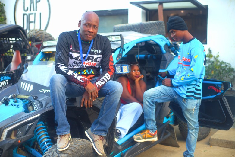 A man sits on a blue-and-black race car before the start of the Ruff Ryders Block Party on March 7, 2026, in Cap-Haïtien. Photo by Onz Chéry / The Haitian Times.