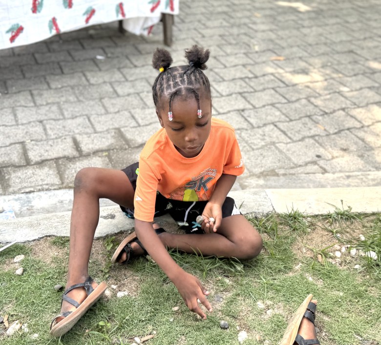 A little girl practices playing jacks with small rocks during “Let’s Play Together” on Saturday, Aug. 15, 2026, at Lycée Anacaona in Léogâne, Haiti. Photo by Daniella Saint-Louis for The Haitian Times.